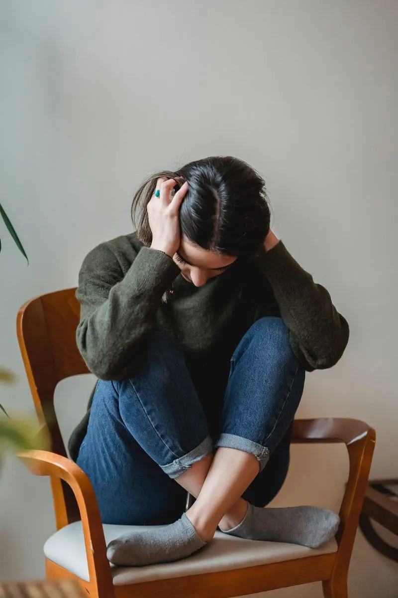 A woman with anxiety is sitting cross-legged on a chair, cradling her head in her hands.