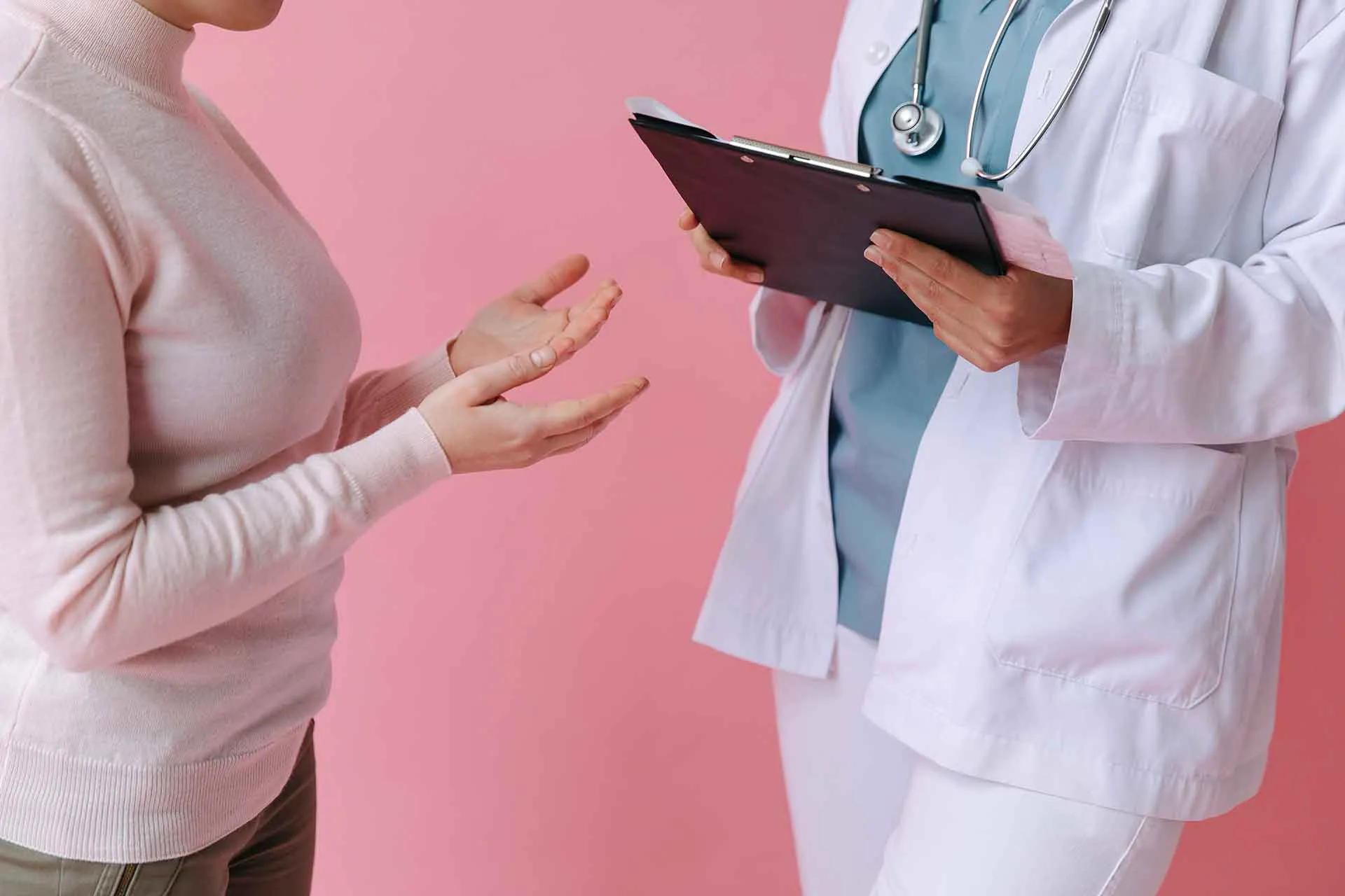 Patient in a pink sweater gesturing while talking to a doctor holding a clipboard