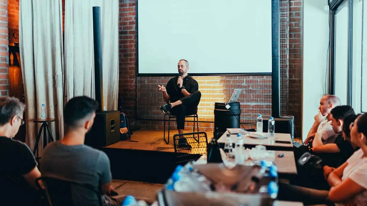 Speaker with a microphone seated on a stage addressing a small audience in a brick-walled venue