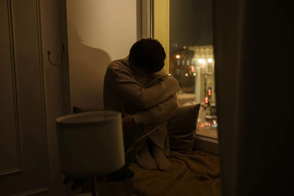 Person hugging their knees on a window seat at night in a dimly lit room